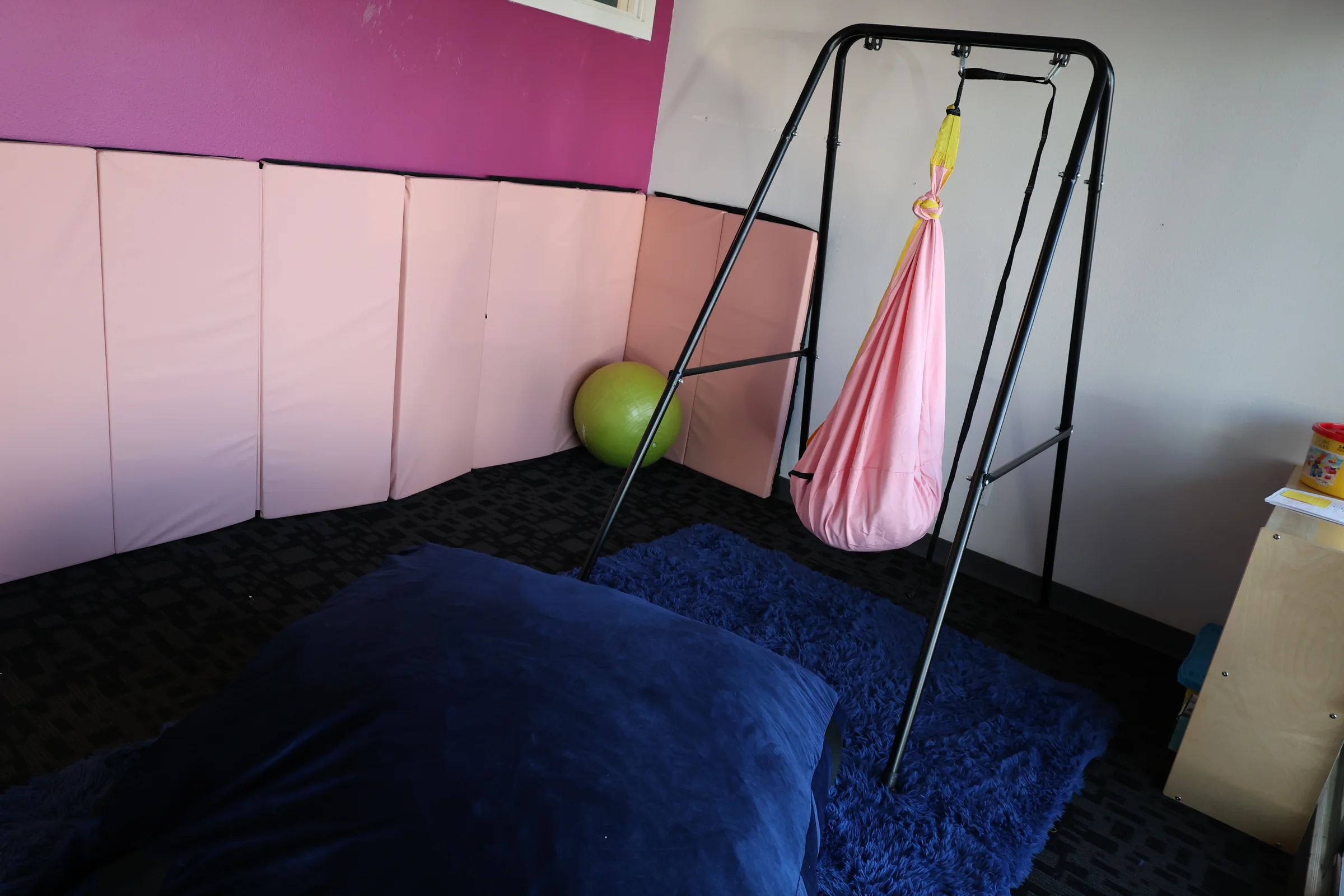 A regulation space with padded wall mats, a hanging fabric swing, a large floor cushion and an exercise ball.